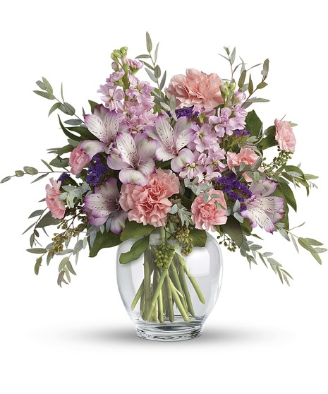  floral arrangement in a clear glass vase featuring soft pink carnations, lavender alstroemeria, delicate purple stock, and lush greenery.